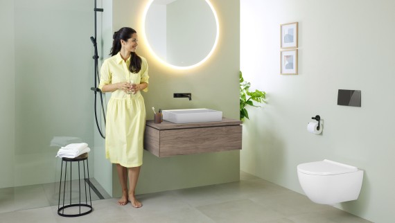Femme en robe jaune devant une salle de bains vert menthe avec meubles et céramiques de salle de bains Geberit et robinets noirs.
