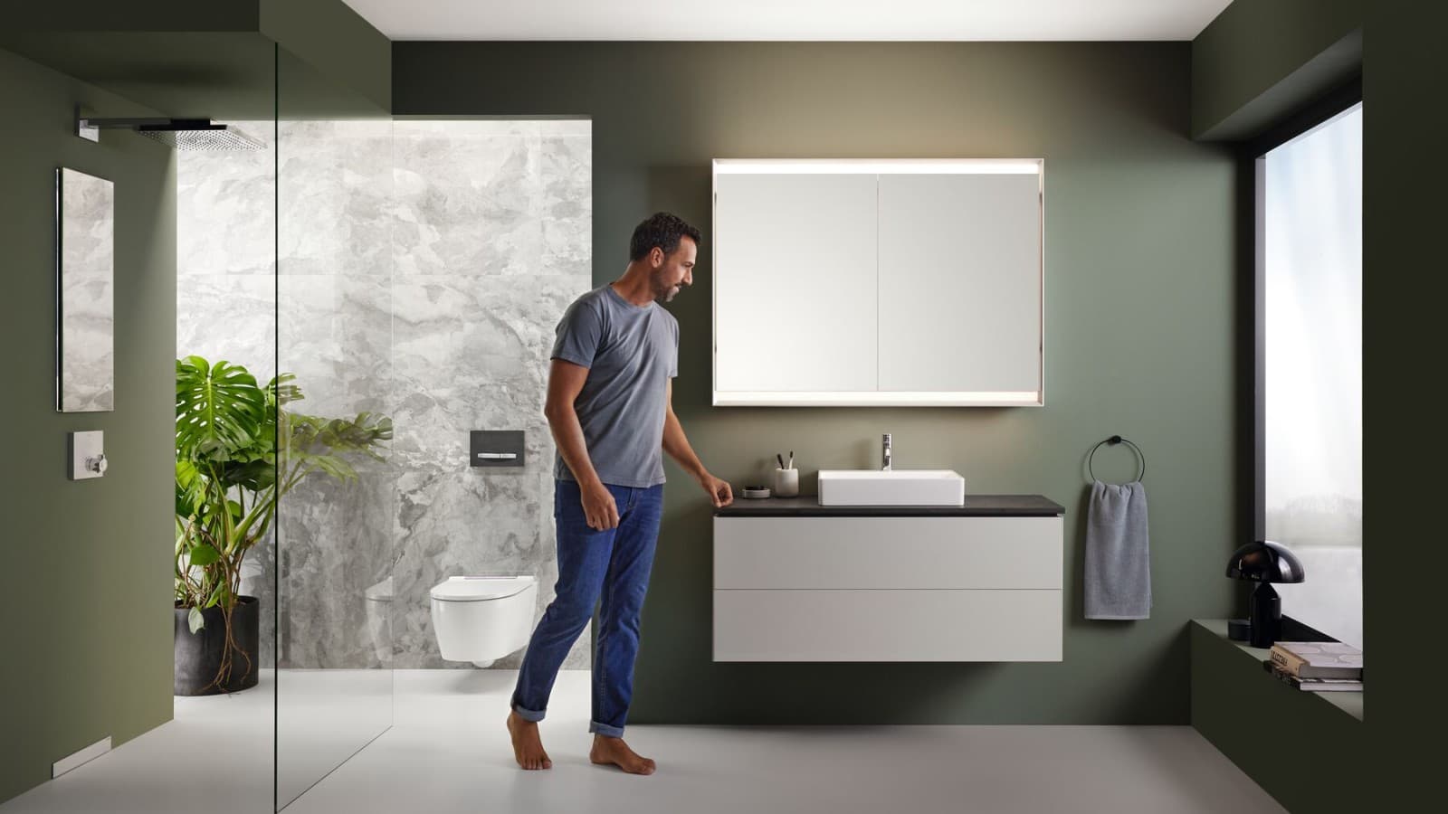 Un homme se tient dans une salle de bains équipée de meubles de haute qualité de la série Geberit One Un homme se tient dans une salle de bains équipée de meubles de haute qualité de la série Geberit One