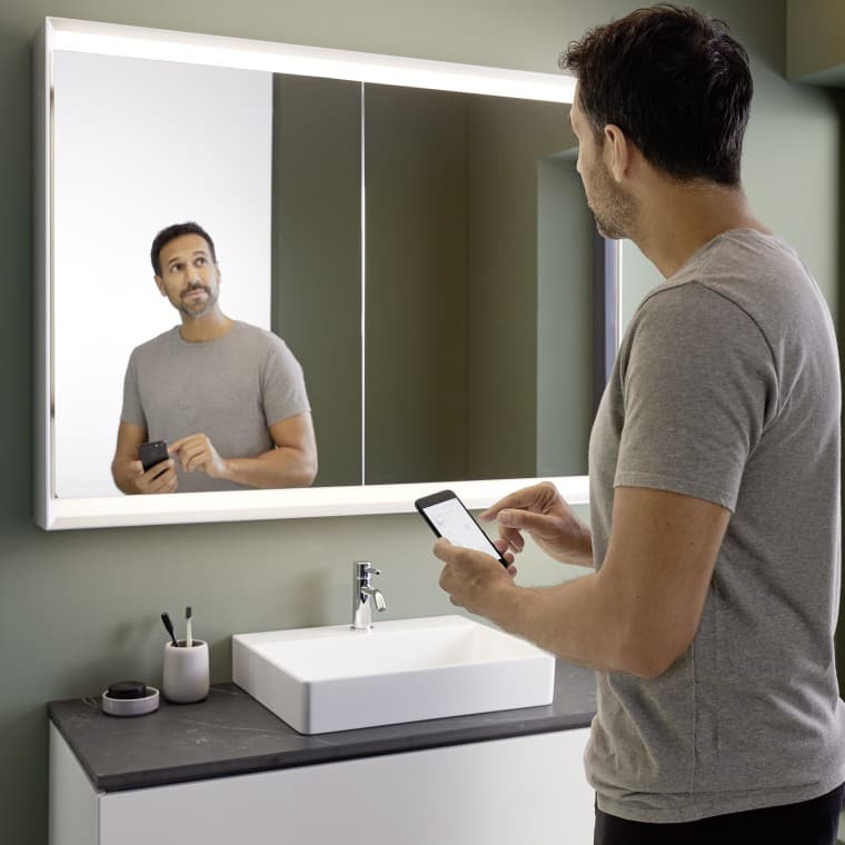 Un homme commande lʼéclairage de lʼarmoire à miroir Geberit ONE avec son smartphone (© Geberit) Un homme commande lʼéclairage de lʼarmoire à miroir Geberit ONE avec son smartphone (© Geberit)